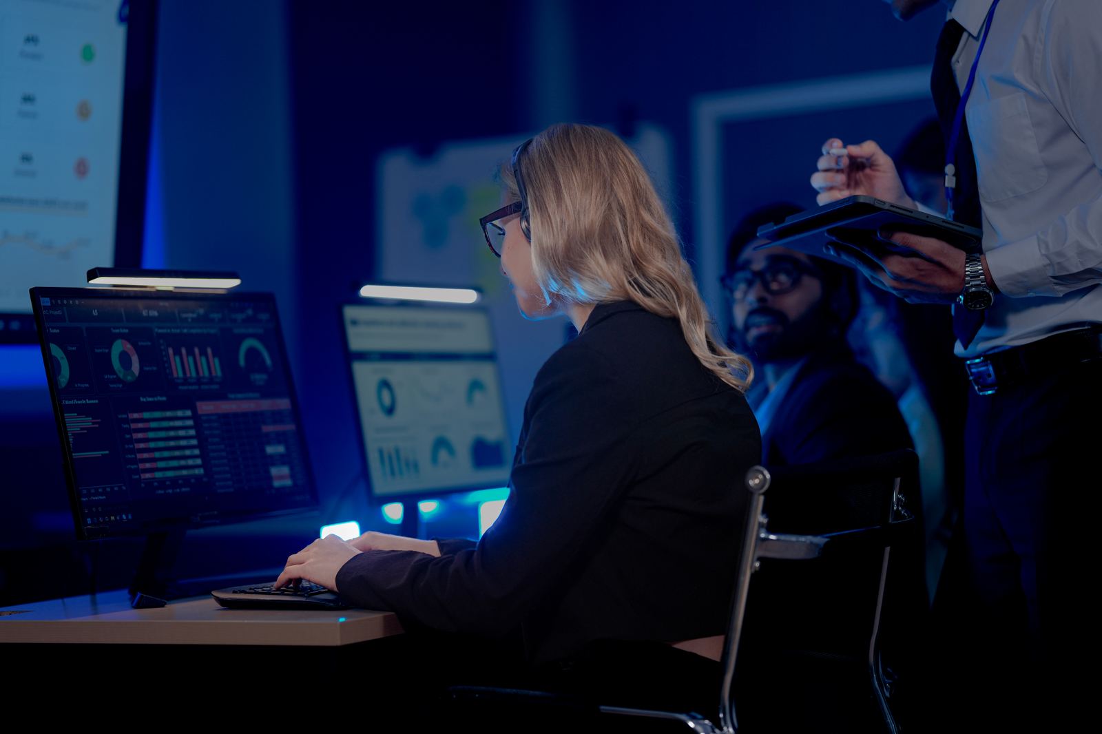 An analyst reviewing live dashboard charts across multiple monitors, colleagues in the background